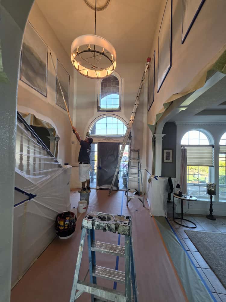 Simply Pro Painting crew member on a ladder painting a tall two-story foyer with floors and furnishings fully protected